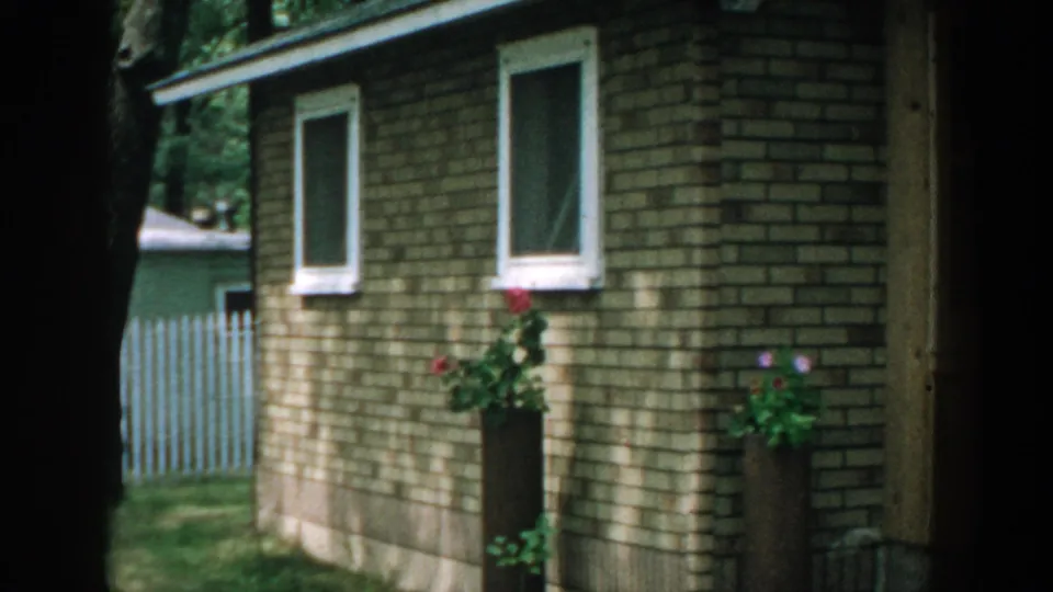 1957 Wisconsin home-movie frame showing a brick house exterior with white windows, potted flowers, lawn, trees, and a fence.