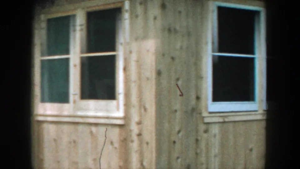1957 Wisconsin home-movie view of a tan wooden sunroom or shed wall with windows, grass, and dark frame edges.