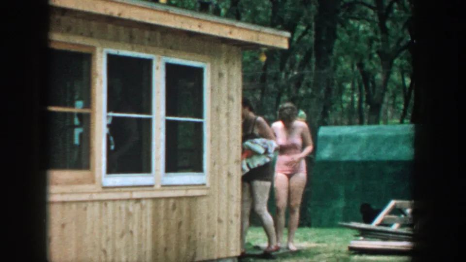 1957 Wisconsin backyard frame showing women in swimsuits near a tan wooden sunroom, with lawn, trees, and outdoor items nearby.