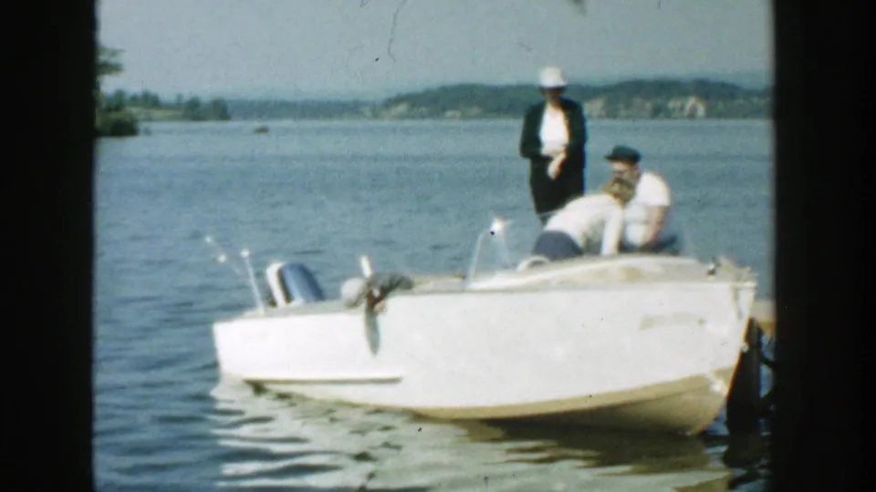 Color 8mm frame from Wisconsin in 1957 showing three family members around a white motorboat on calm lake water.