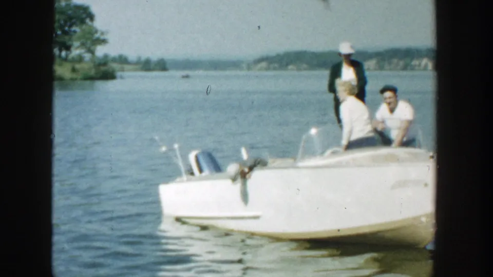 Color home-movie frame from 1957 Wisconsin showing a family beside a white motorboat on a sunny lake.