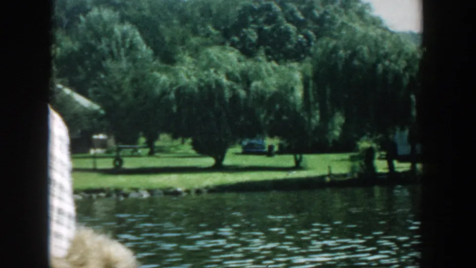 1957 Wisconsin lakeshore view from a moving boat, showing willow trees, grass, and water with a partial passenger at left.