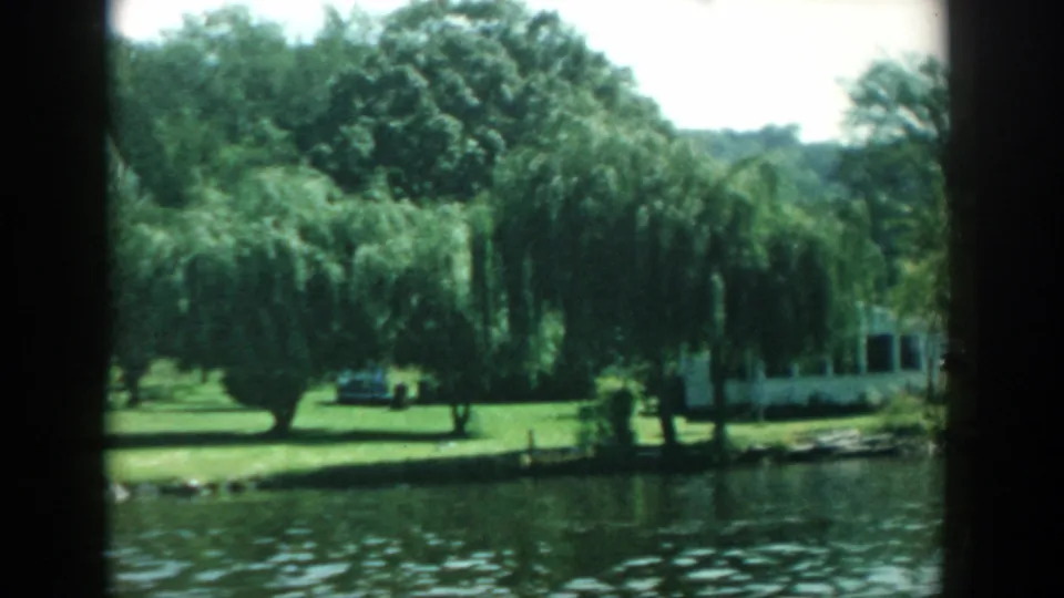 Wisconsin lake shoreline in 1957 home-movie footage, with willow trees, grass, water, and a light-colored lakeside building.