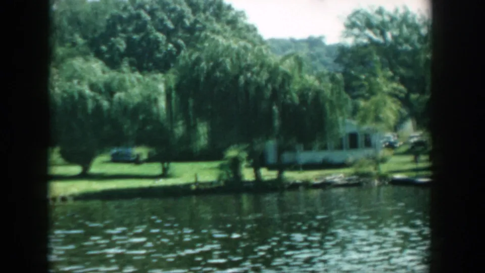 1957 Wisconsin lakeside park scene with rippling water, willow trees, maintained grass, a pale building, and mid-century cars in the background.