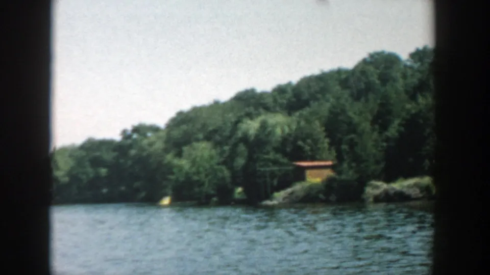 1957 Wisconsin 8mm frame with open lake water, a wooded shoreline, a willow tree, and a small yellow shoreline building.