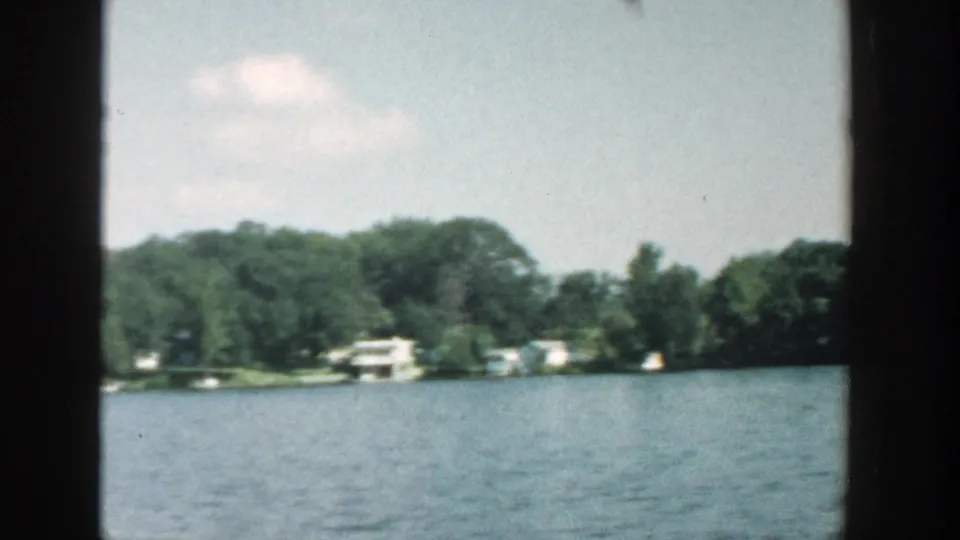 1957 Wisconsin lake view with tree-lined shore, scattered cottages, and blue water filmed from a moving vantage point.