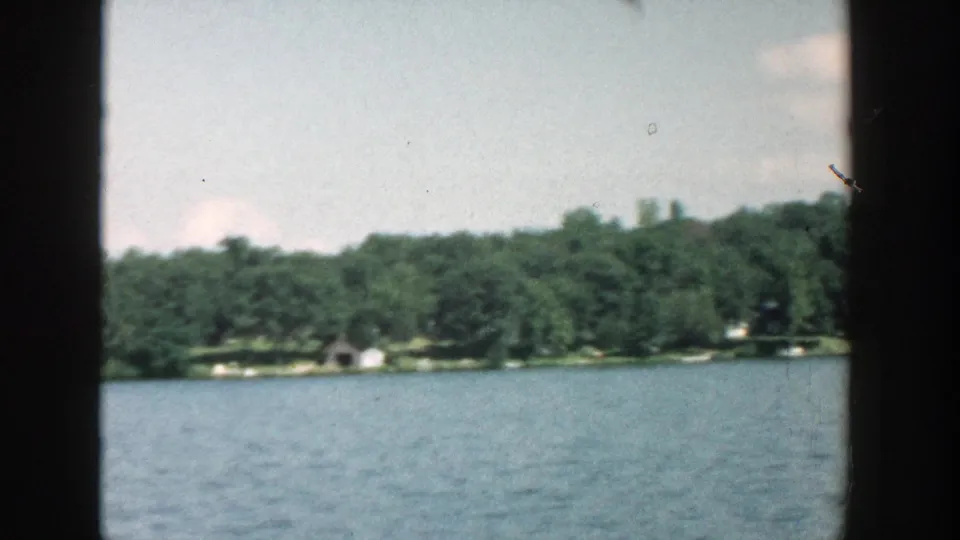 Wisconsin 1957 home-movie frame showing lake water, dense green trees, and small shoreline buildings under a pale sky.
