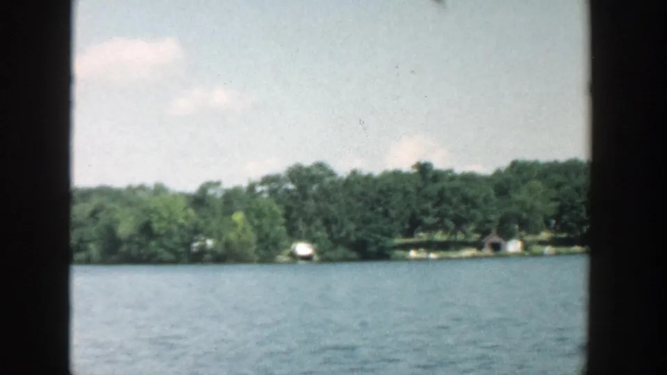 1957 Wisconsin lakeside view with calm water, dense shoreline trees, and small distant structures framed by 8mm film texture.