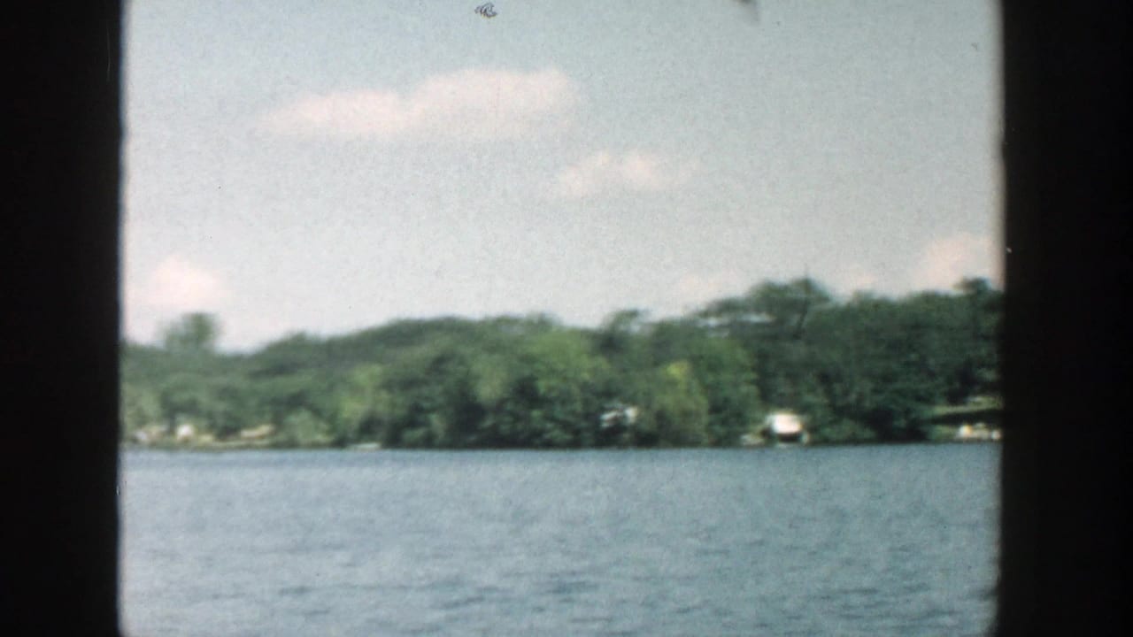 Calm lake water with a wooded shoreline and distant houses seen through dark window framing.