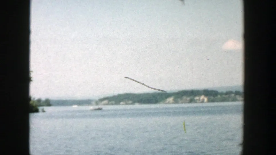 1957 Wisconsin lake scene from 8mm film, showing open water, a distant shoreline, faint boat activity, pale sky, and visible film scratches.