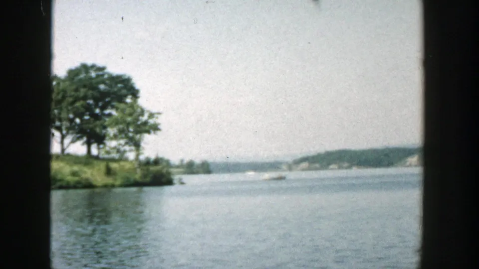 1957 Wisconsin 8mm frame showing a small tree-covered island or point, calm lake water, distant shoreline, and pale summer sky.