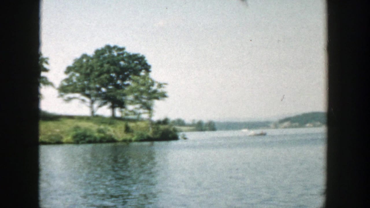 1957 Wisconsin 8mm frame showing calm lake water, a grassy wooded island, distant shoreline, and dark contact-frame edges.