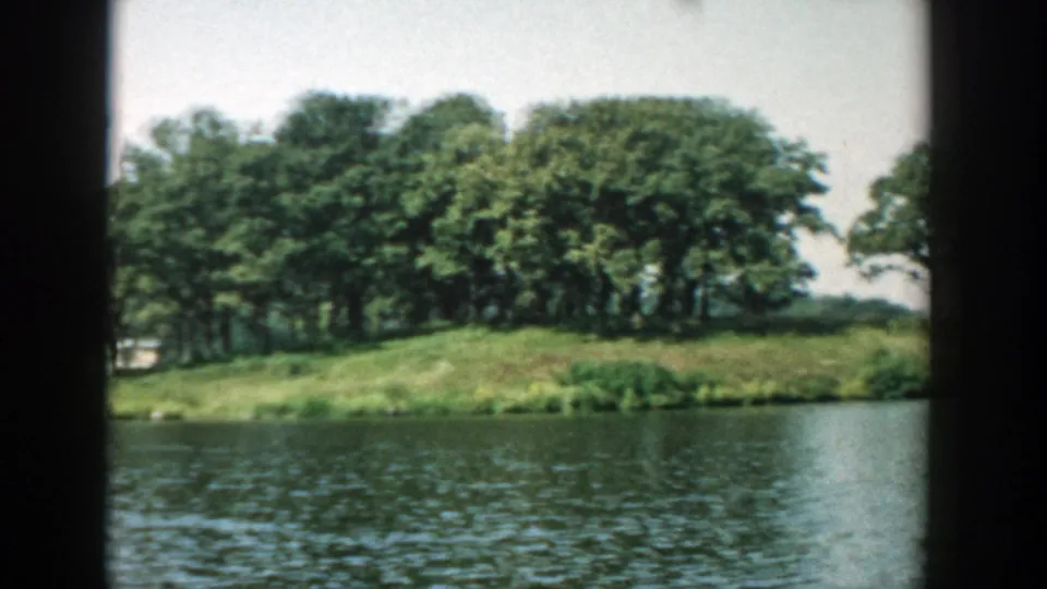 1957 Wisconsin 8mm frame with rippled lake water, a low grassy bank, and dense trees on a small wooded island or shoreline.