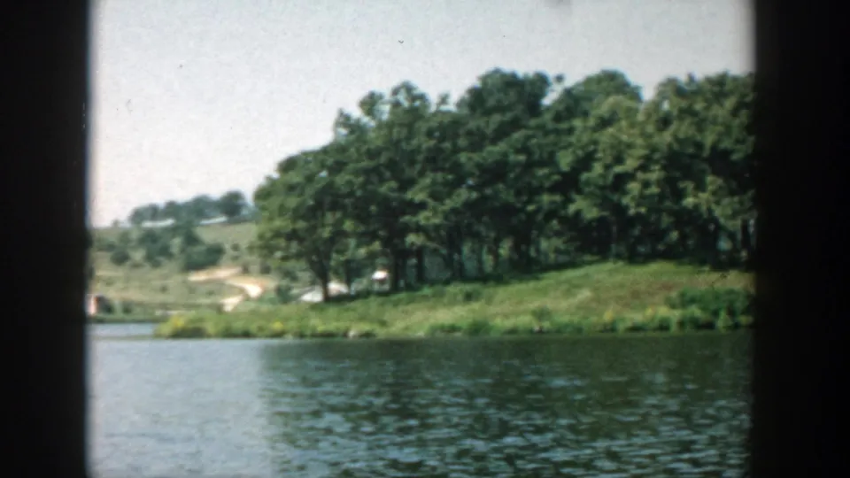 1957 Wisconsin lake scene showing water in the foreground, a wooded grassy bank, distant hillside, and soft 8mm exposure.