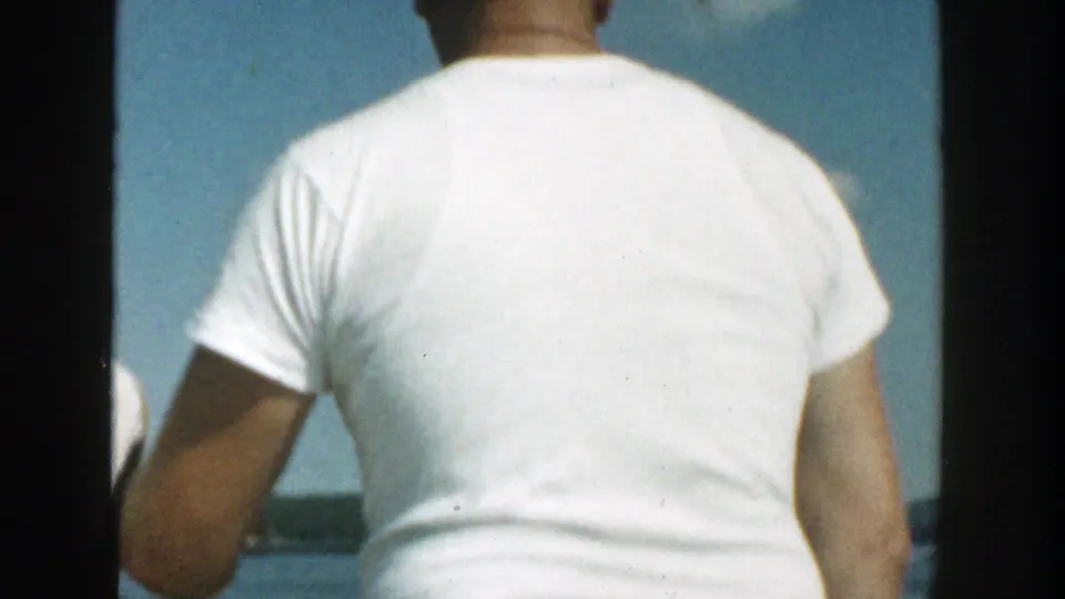 1957 Wisconsin home-movie frame of an adult seen from behind on a boat, facing open water with a distant dark shoreline.