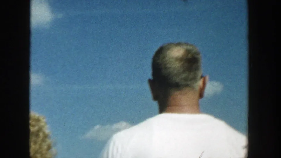 1957 Wisconsin 8mm frame showing an adult from behind, cropped close against a bright sky with small clouds and partial water view.