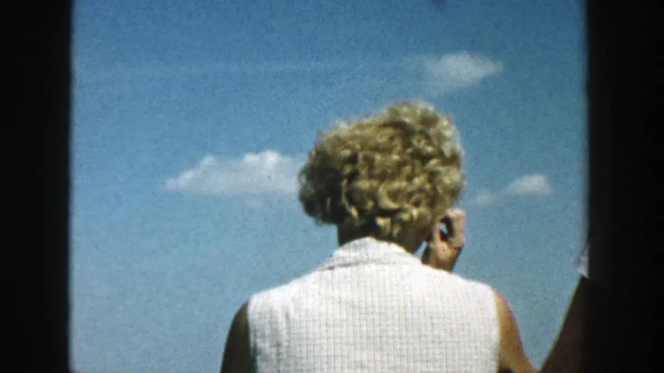 1957 Wisconsin home-movie frame of a blonde woman on a boat, seen from behind with lake and clouds beyond her.