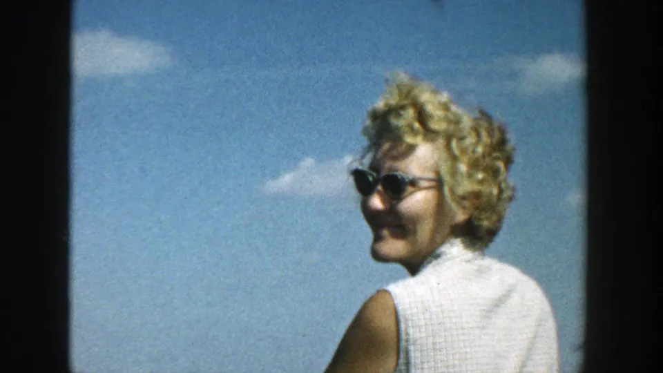 A woman in dark sunglasses and a white sleeveless top turns toward the camera in 1957 Wisconsin outdoor footage.
