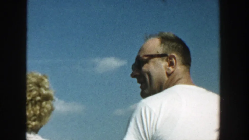 A man in glasses and a white T-shirt stands beside a partly visible woman under a blue sky in Wisconsin home-movie footage.