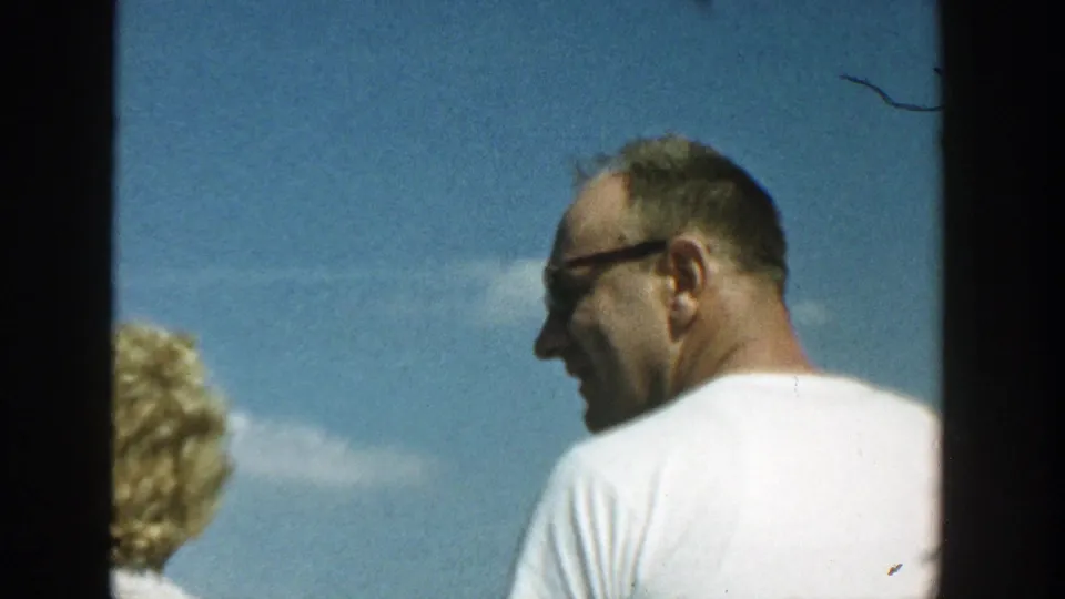 A man in glasses and a white T-shirt looks sideways outdoors, with part of the woman visible at frame edge.
