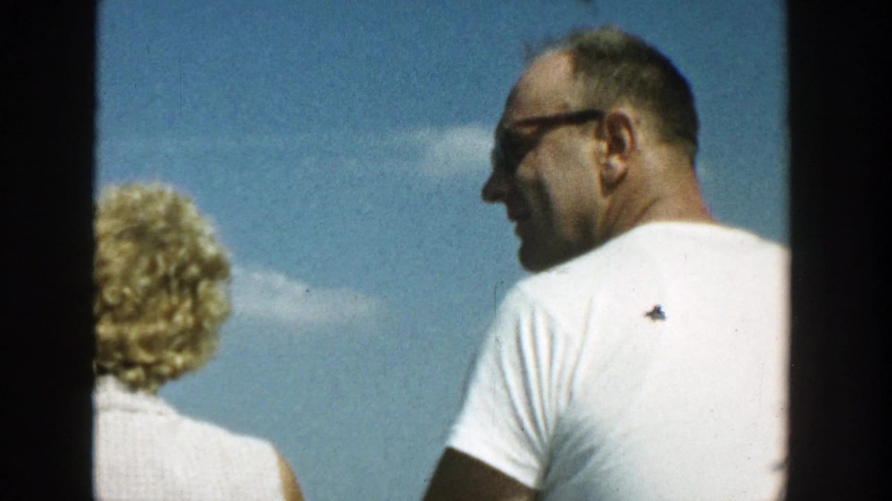 Man in sunglasses and a white T-shirt stands on a boat beside a blonde woman under blue sky.