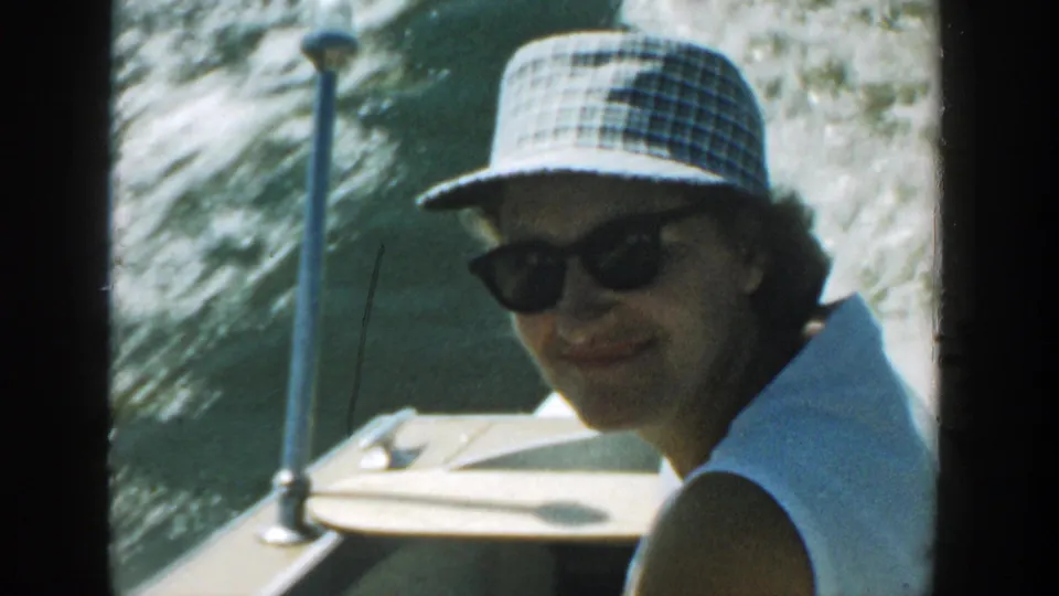 Woman wearing a plaid bucket hat and sunglasses smiles toward the camera on a moving boat in Wisconsin, 1957.