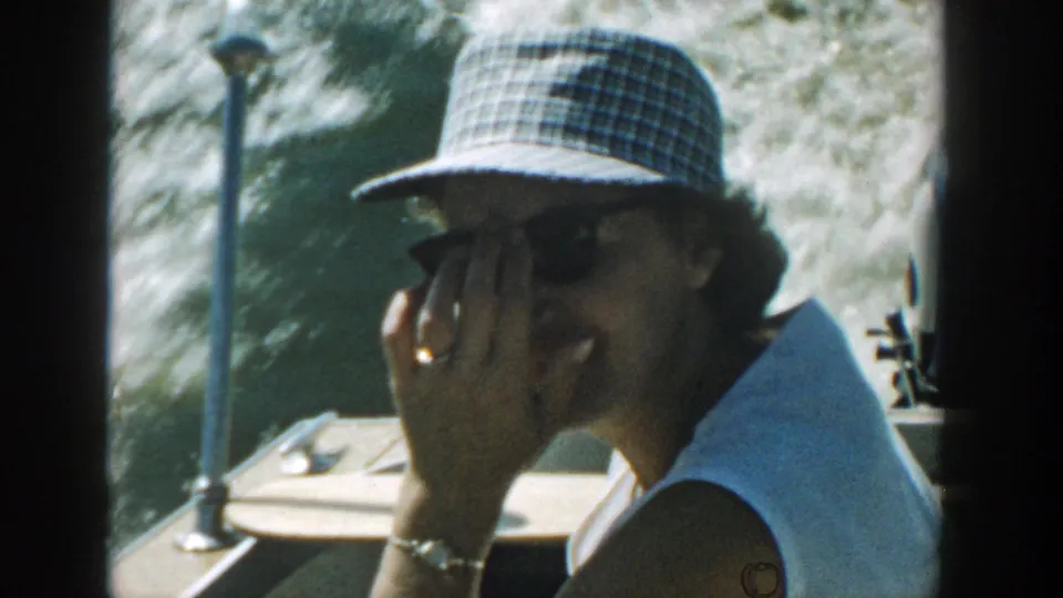 Woman in sunglasses and a plaid hat touches her face while seated on a moving boat in Wisconsin, 1957.