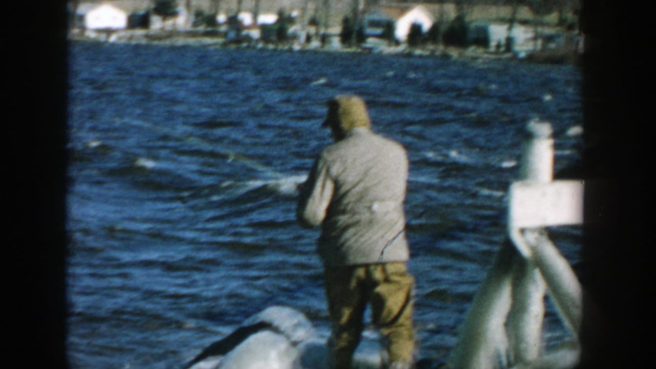 Man in a hooded jacket fishes from rocks beside choppy water.