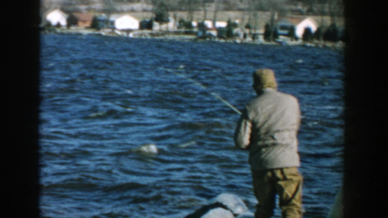 Man fishing from rocks with choppy water and houses in the distance.