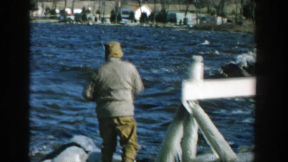 From a handheld 8mm view, the man reels with his back to the camera beside choppy water, rocks, and a white railing.