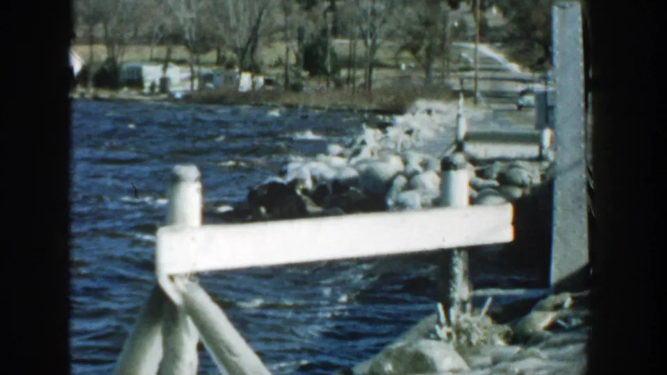 The angler has moved out of view, leaving rocks, rough water, a white rail, and the opposite Wisconsin shoreline in frame.