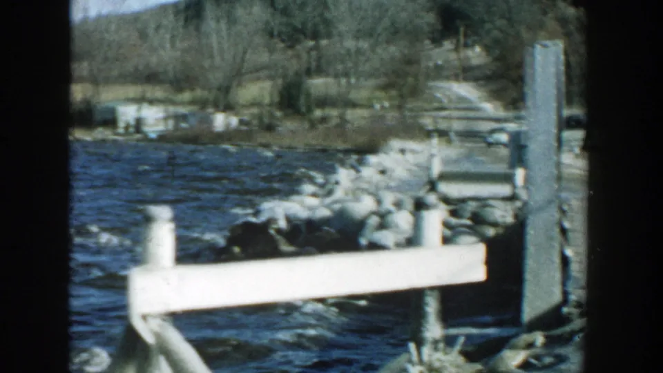 1957 Wisconsin 8mm frame showing a white dock rail, wind-ruffled lake water, rocks, bare trees, and a distant shoreline.