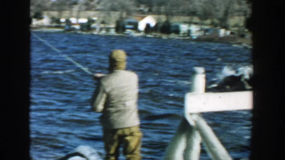 A man in a cap stands near a white dock rail in Wisconsin, casting a fishing line over wind-ruffled lake water.