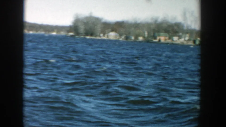 1957 Wisconsin home-movie frame with rippled blue water, low distant shore, small houses, and bare trees.