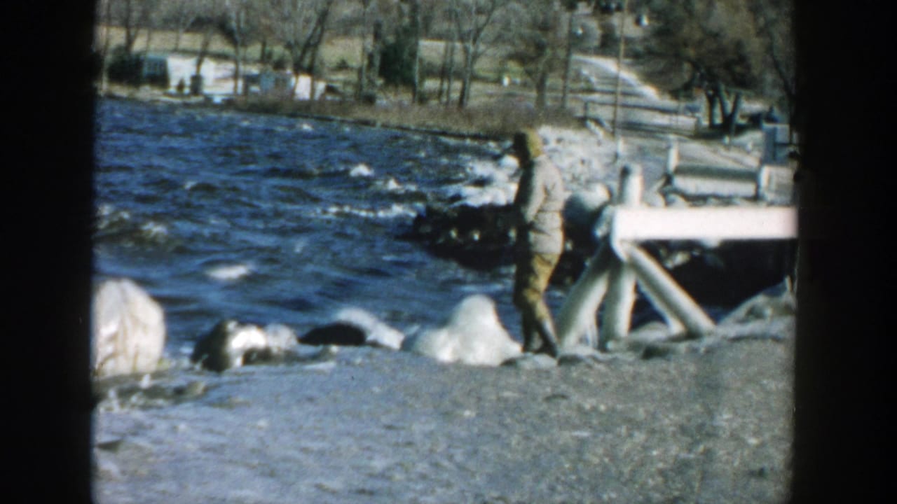 Man in a light jacket stands on a rocky lakeshore near choppy water and small buildings.