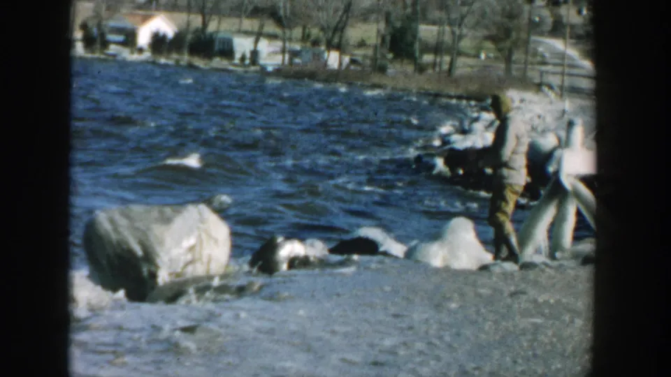 1957 Wisconsin 8mm frame showing a man beside large shoreline rocks with lake water, trees, and buildings beyond.