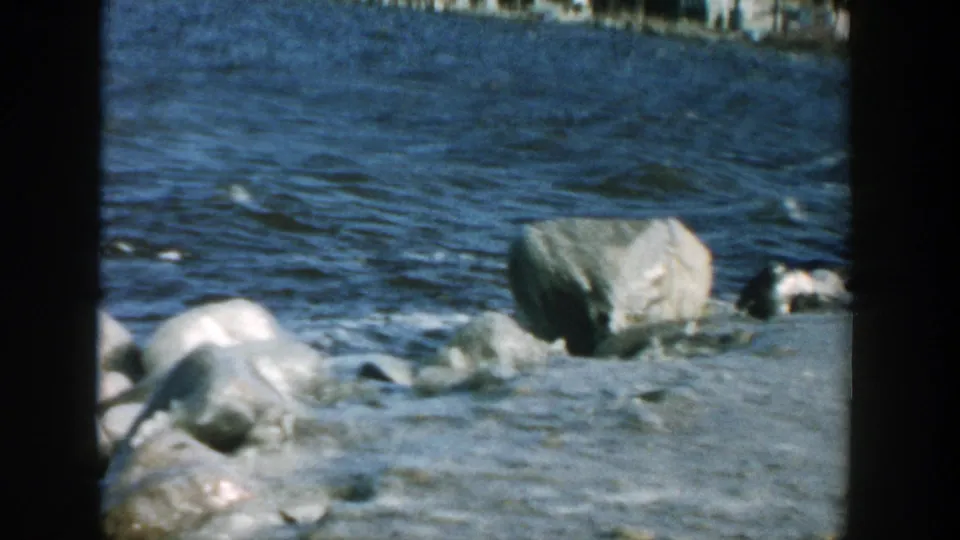 1957 Wisconsin lakeshore detail with choppy blue water washing near large pale rocks and a sloped bank.
