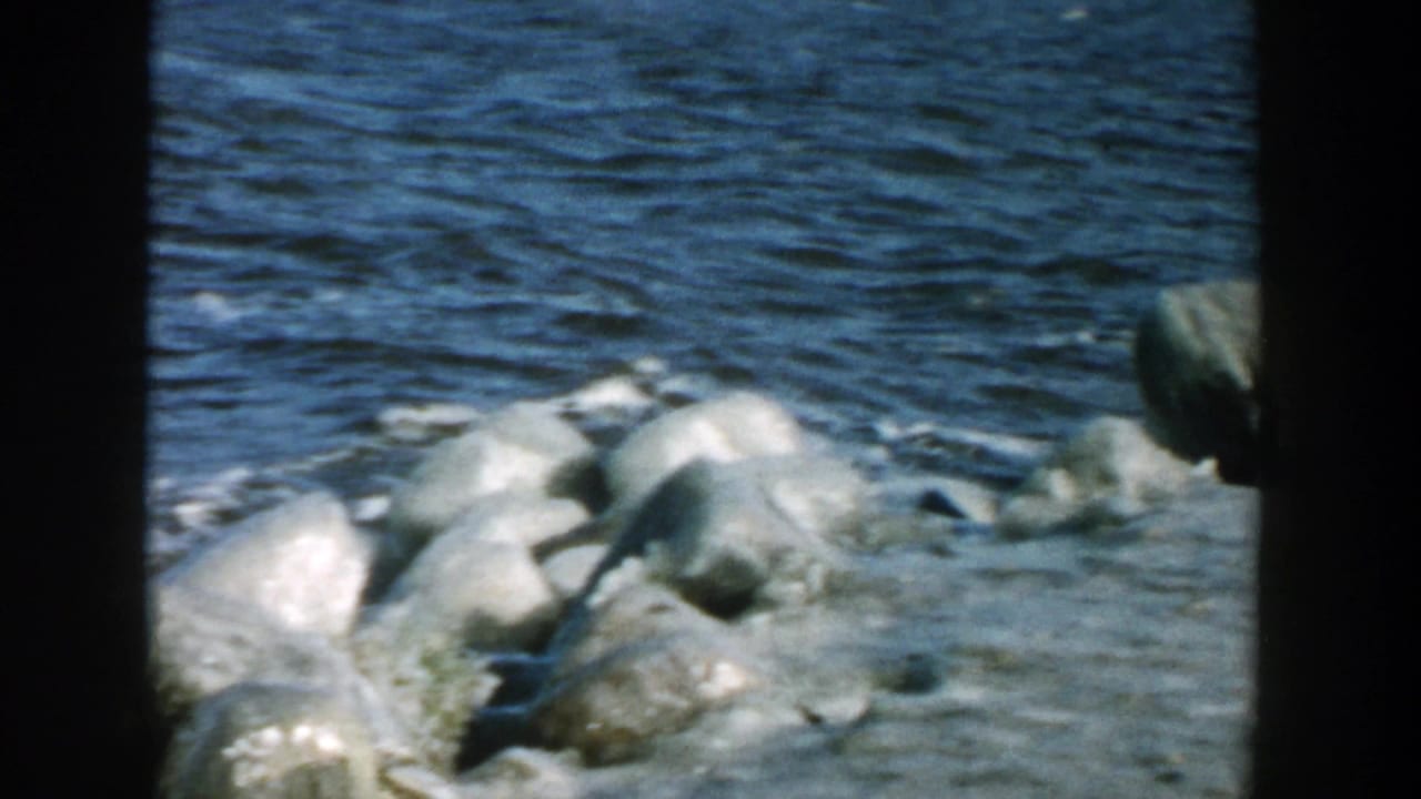 1957 Wisconsin 8mm frame showing rounded shoreline rocks, pale ice, and fast-moving dark river water.