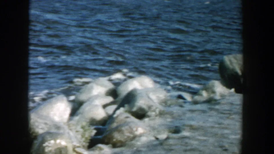 1957 Wisconsin 8mm frame showing rounded shoreline rocks, pale ice, and fast-moving dark river water.