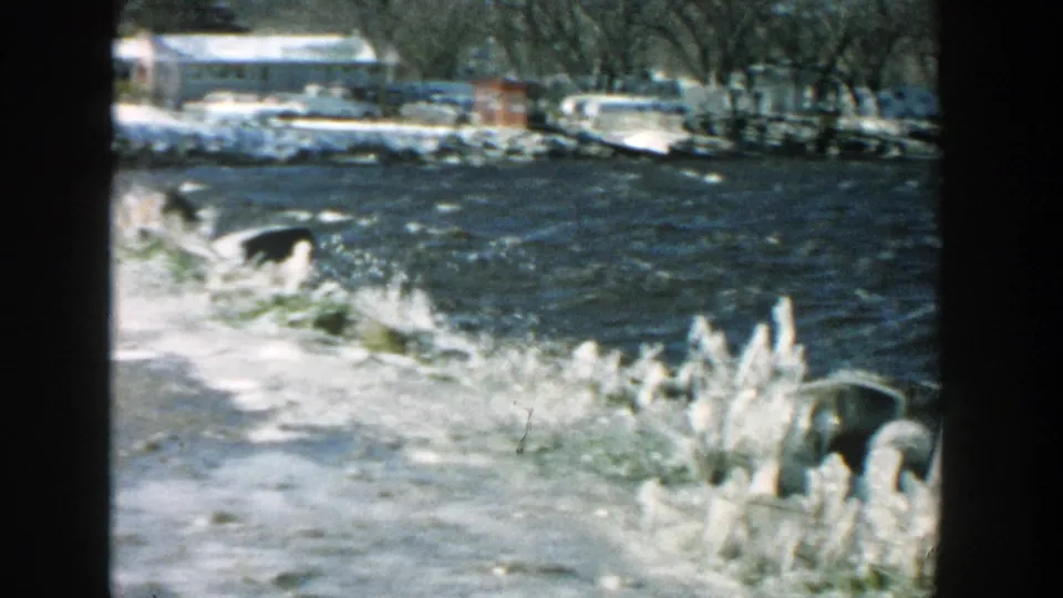 1957 Wisconsin 8mm frame showing icy riverbank foreground with a distant residential area and parked cars beyond the water.