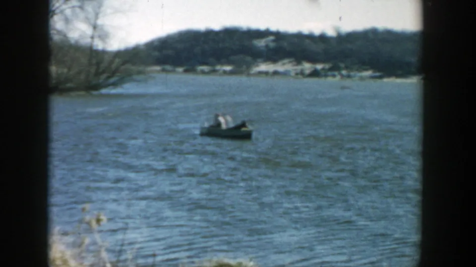 Small boat with distant people on a Wisconsin river, with a wooded hill and shoreline structures in the background.
