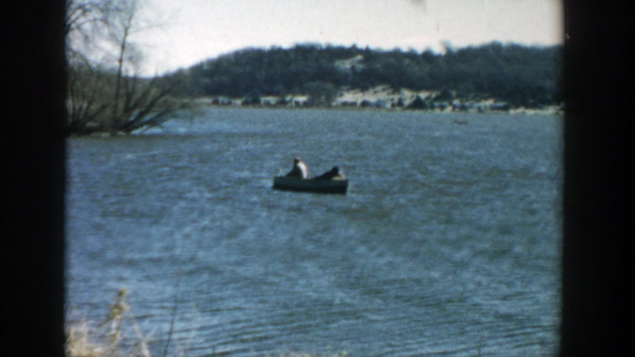 1957 Wisconsin home-movie frame of two people in a small motorboat on open water, with bare trees and low hills in the distance.