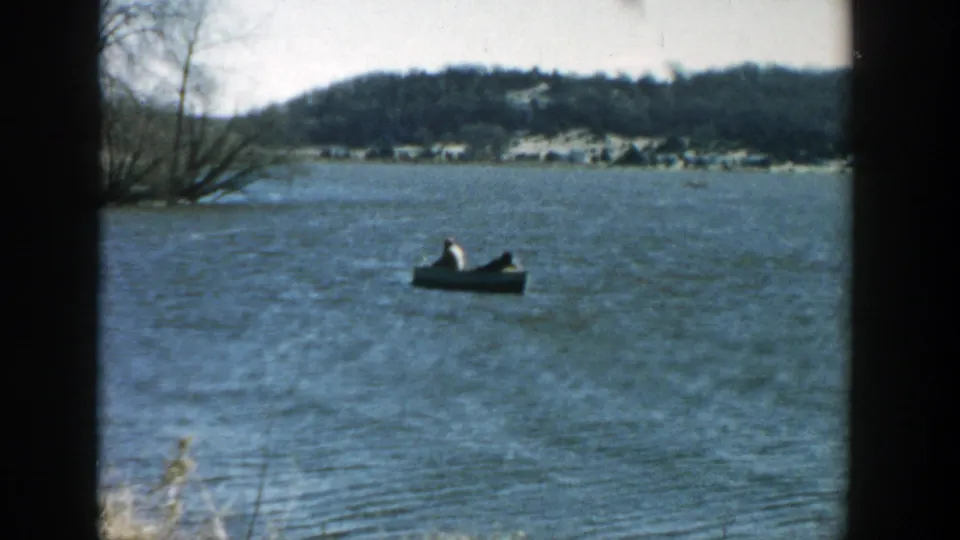 1957 Wisconsin home-movie frame of two people in a small motorboat on open water, with bare trees and low hills in the distance.