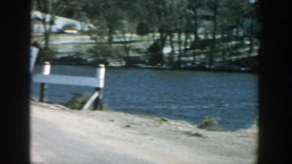 1957 Wisconsin frame showing a grey wooden bridge or roadside railing beside a lake or river, with trees and shoreline in the background.
