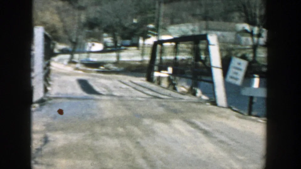 1957 Wisconsin home-movie view down a road over a grey wooden bridge, with water at both sides, trees, and an unreadable white sign.