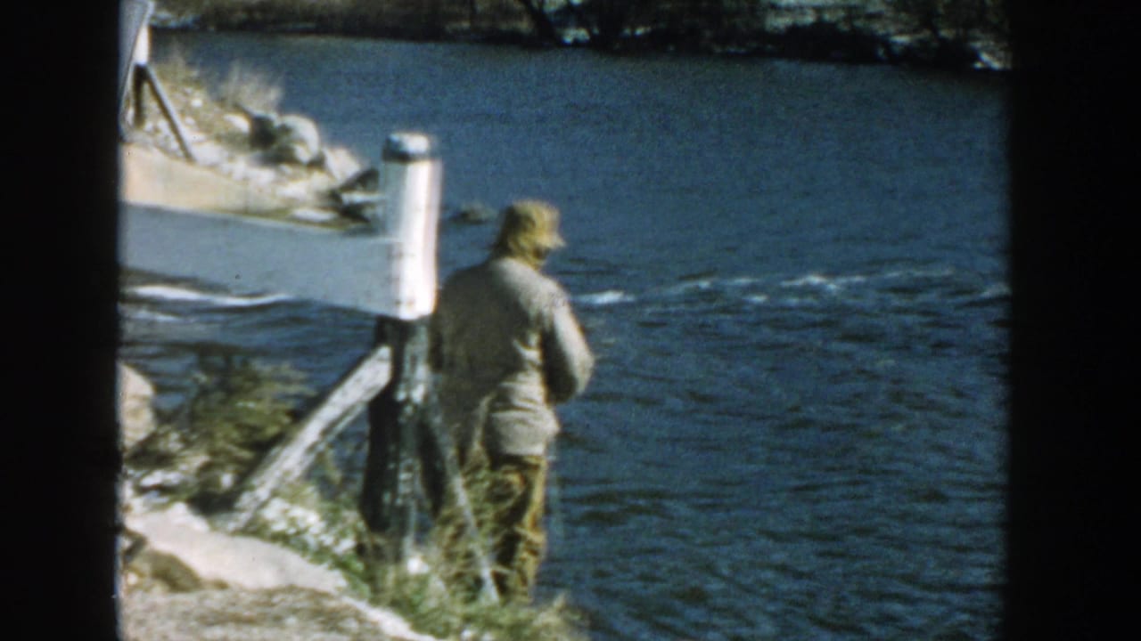 1957 Wisconsin home-movie frame showing a man with a fishing rod beside a river near bridge supports and a white sign.
