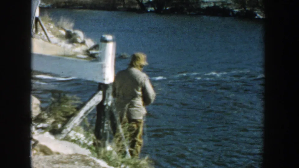 1957 Wisconsin home-movie frame showing a man with a fishing rod beside a river near bridge supports and a white sign.