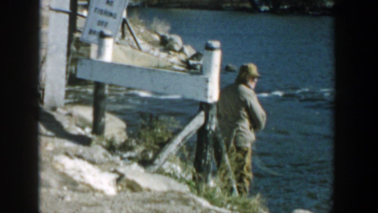 Man fishes beside a river with bridge railing and sign above the bank.