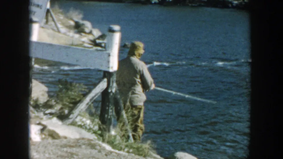 A mid-century home-movie view shows the fisherman at the riverbank with his rod extending over the water near a bridge.