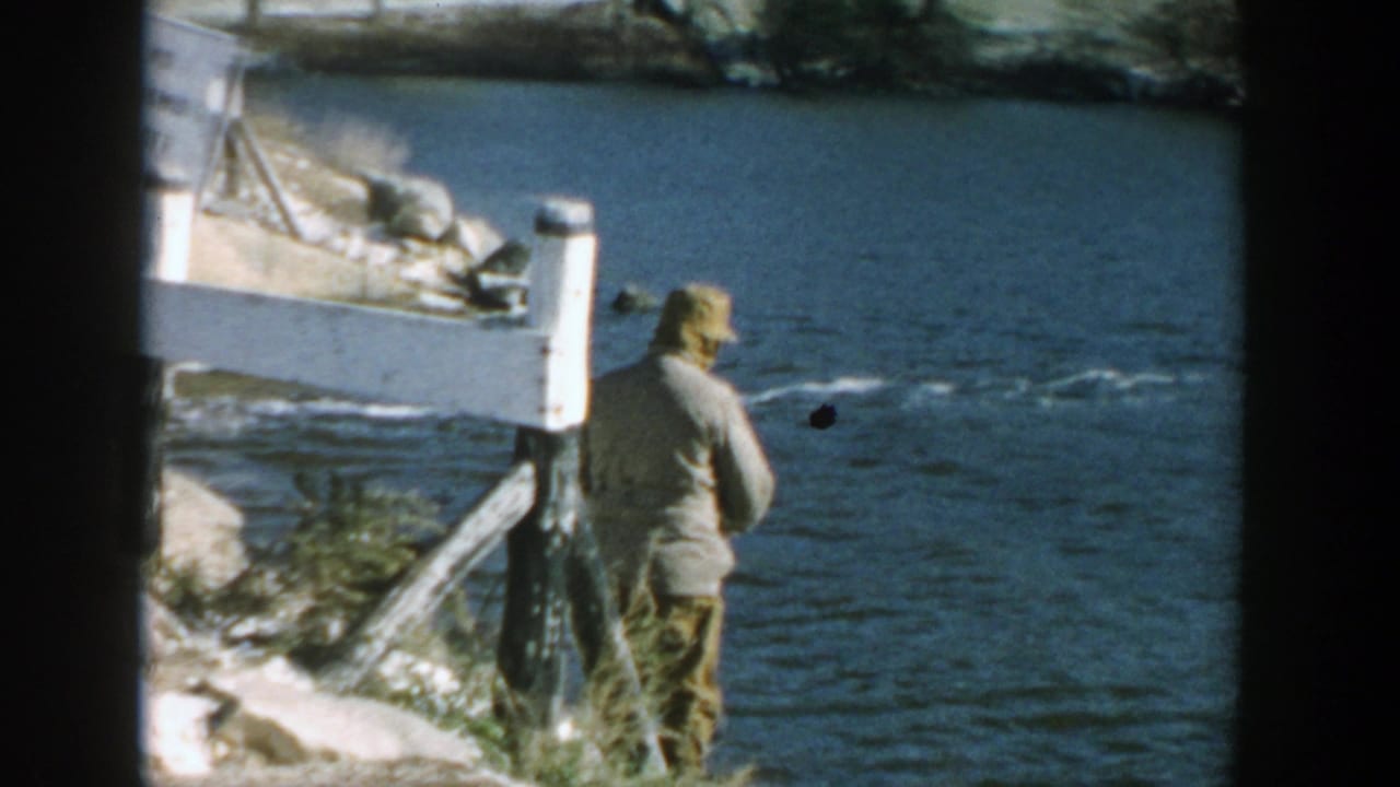 In 1957 Wisconsin home-movie footage, a man in a hat fishes from a rocky riverbank beside rippling water.